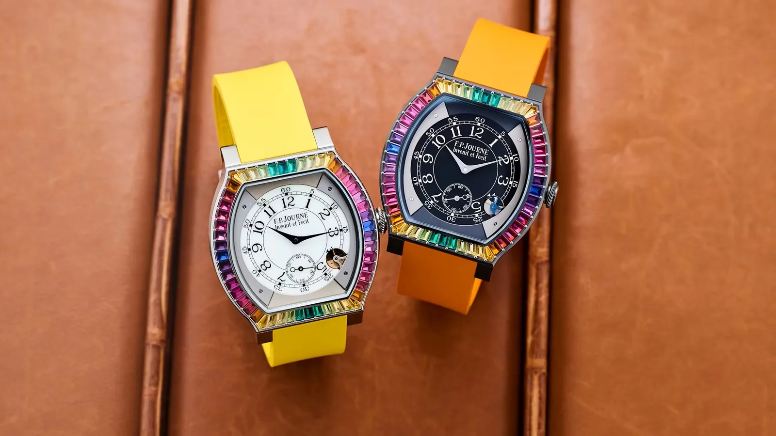 Two F.P. Journe Élégante watches with rainbow baguette-set bezels, one in titanium on a yellow rubber strap and one in Titalyt® on orange, lying on tan leather