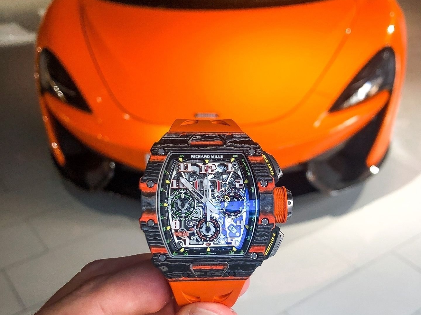 A Richard Mille RM 11-03 McLaren in Carbon TPT and orange Quartz TPT on an orange rubber strap, held up in front of an orange McLaren car